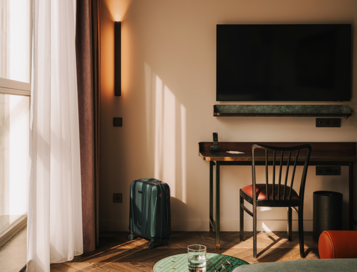 Pokój hotelowy z drewnianą podłogą, biurkiem, telewizorem na ścianie i walizką przy zasłoniętym świetle.