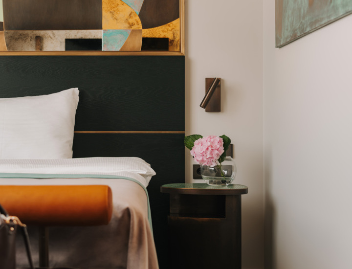 Nowoczesny pokój hotelowy z łóżkiem, abstrakcyjną sztuką nad zagłówkiem i bukietem różowych kwiatów na eleganckim stoliku.