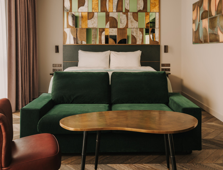Elegancki pokój hotelowy z podwójnym łóżkiem, zieloną sofą, nowoczesnym stolikiem i abstrakcyjną dekoracją ściany.