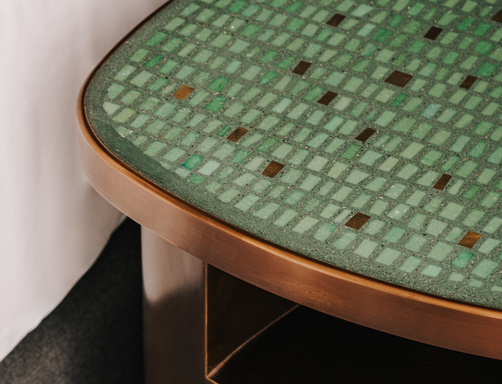 Stolik nocny z mozaikowym blatem w zielonych odcieniach i mosiężną ramą obok łóżka z białą pościelą
