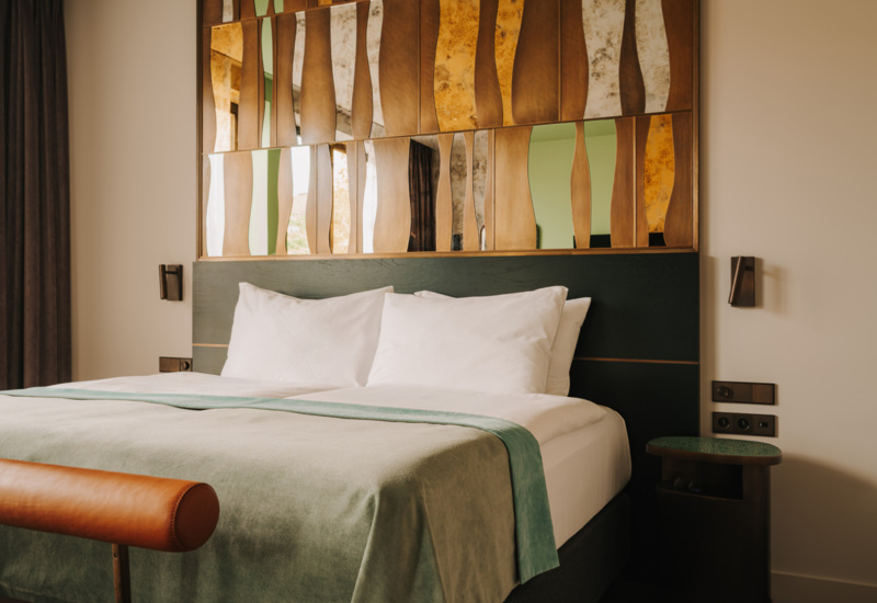 Nowoczesny pokój hotelowy z dużym łóżkiem, białą pościelą, zielonym przebraniem i dekoracyjną ścianą z lustrzanymi elementami.