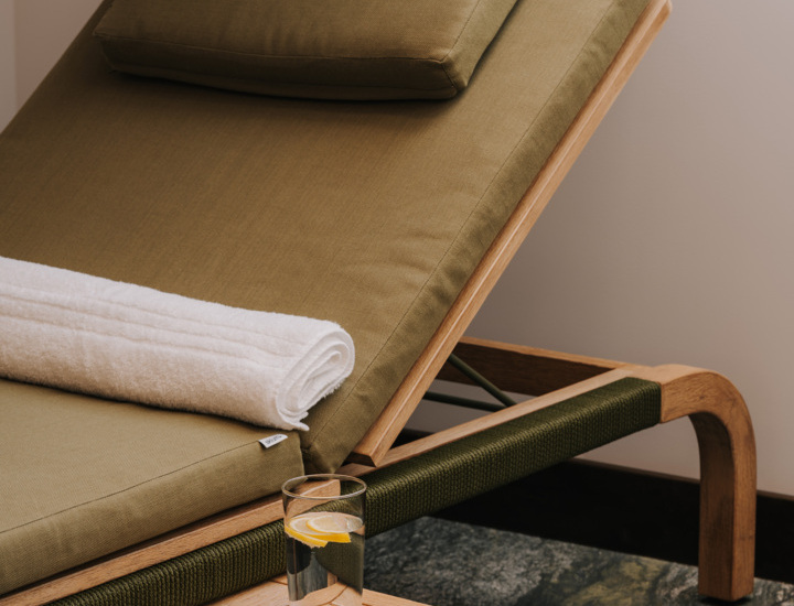 Leżak w strefie spa z zielonym materacem, białym ręcznikiem i szklanką wody z cytryną na drewnianym stoliku.