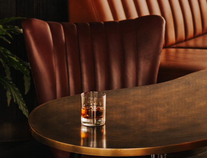 Szklanka z drinkiem na eleganckim stołku przy brązowym, pikowanym fotelu i kanapie w stylowym hotelowym barze.