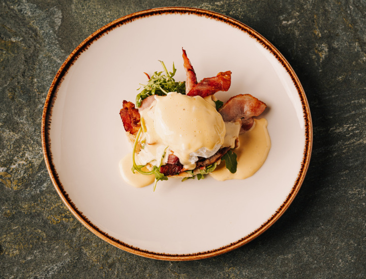 Potrawa na białym talerzu z bekonem, jajkiem w sosie holenderskim i świeżą rukolą na kamiennym stole