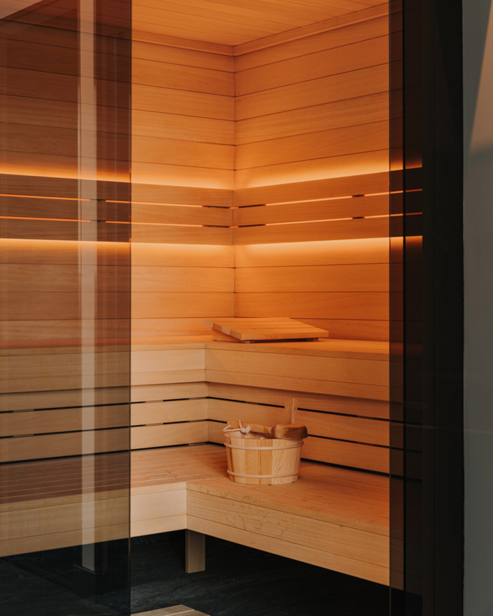 Nowoczesna sauna hotelowa wykończona jasnym drewnem, z ciepłym oświetleniem i drewnianym wiadrem z chochlą dla relaksu gości.