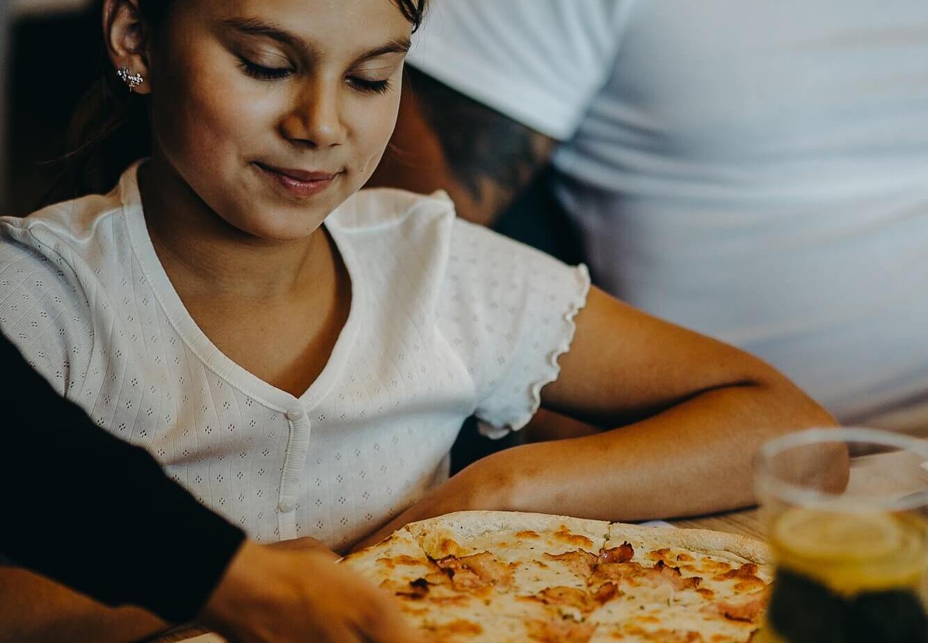 Dziewczynka z zamkniętymi oczami czeka na pizzę podawaną w restauracji, przy stole z napojem i szklanką wody.