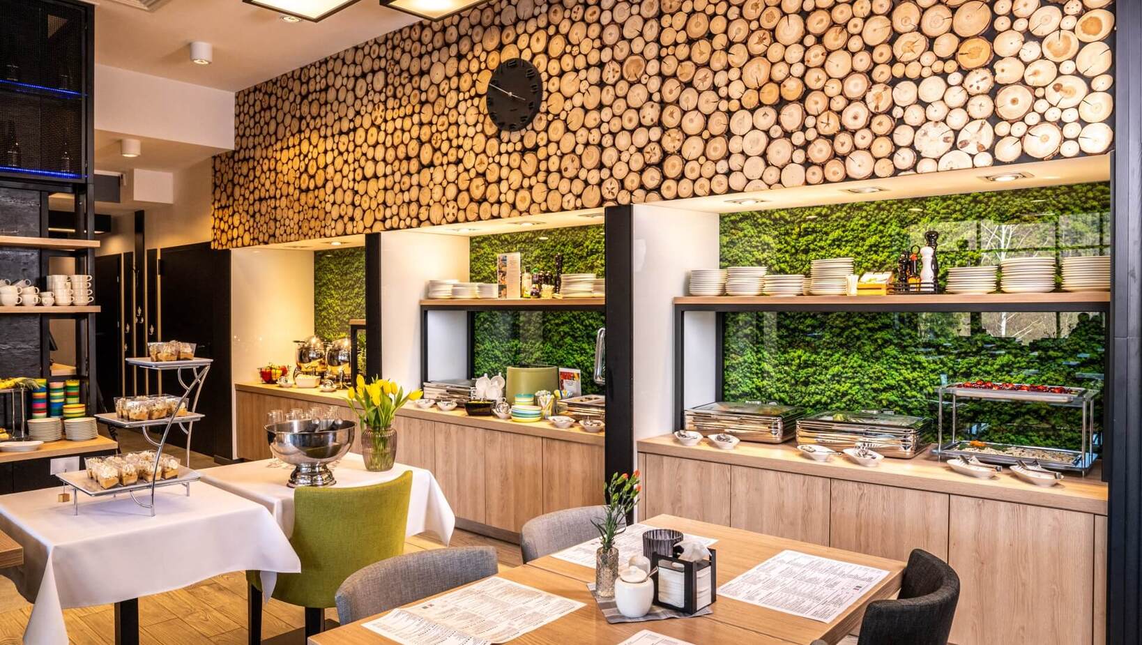 Jasna jadalnia hotelowa z bufetem śniadaniowym, drewnianą ścianą z przekrojami pni i zieloną roślinną ścianą.