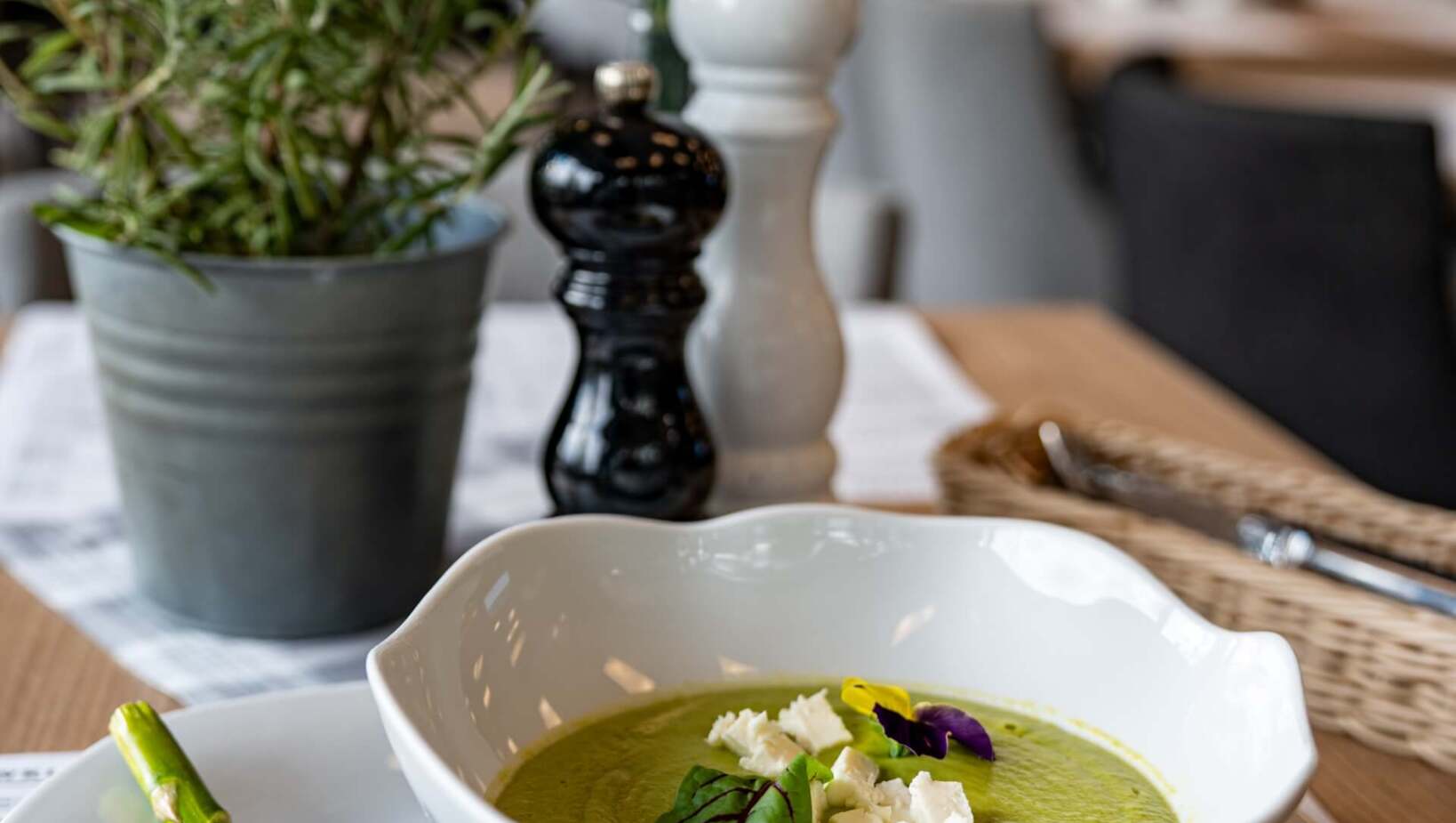 Kremowa zupa z zielonych warzyw z serem i jadalnymi kwiatami, podana z szparagiem na eleganckim stole restauracyjnym.