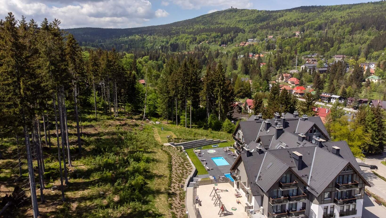 Hotel z tradycyjną górską architekturą, basenem, placem zabaw i widokiem na zielone lasy oraz okoliczne wzgórza.