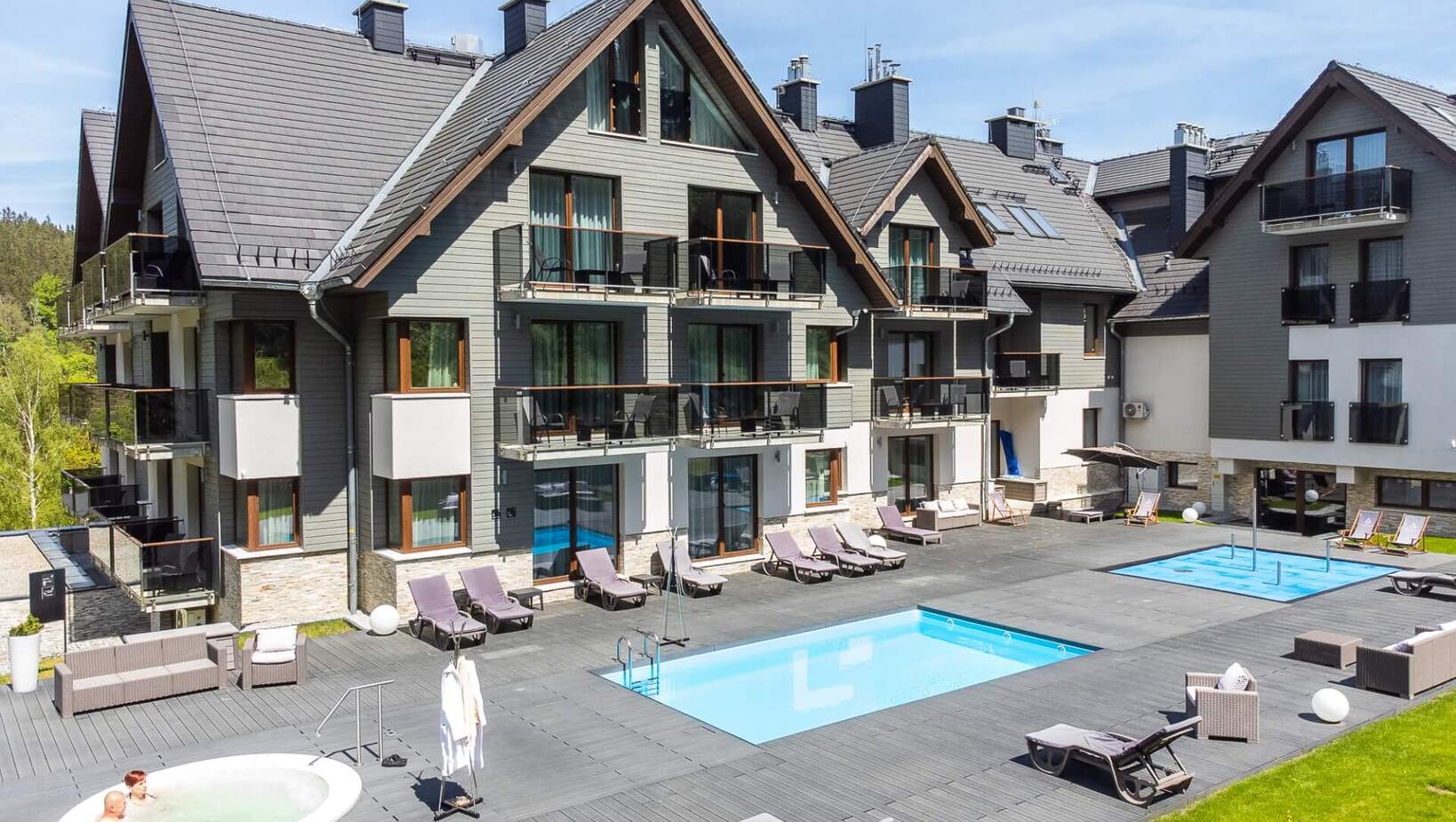 Nowoczesny hotel z basenem, jacuzzi, leżakami i balkonami, otoczony zielenią w spokojnej okolicy.