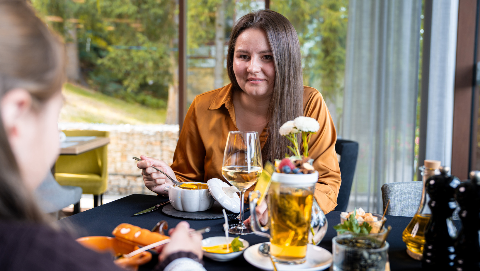 Frau im orangefarbenen Oberteil isst Suppe am elegant gedeckten Tisch; Wein, Vorspeisen und große Fenster mit Gartenblick