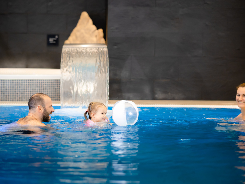 Rodzina bawiąca się z piłką w basenie hotelowym z wodospadem z białymi kafelkami i ciemną ścianą