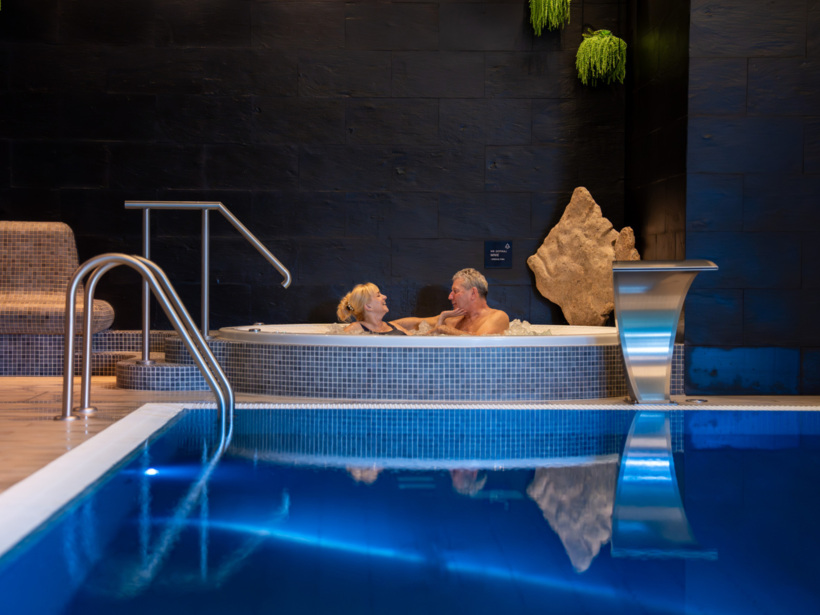 Para relaksująca się w jacuzzi w stylowym spa z basenem i nowoczesnym wnętrzem z czarną ścianą i zielonymi roślinami.