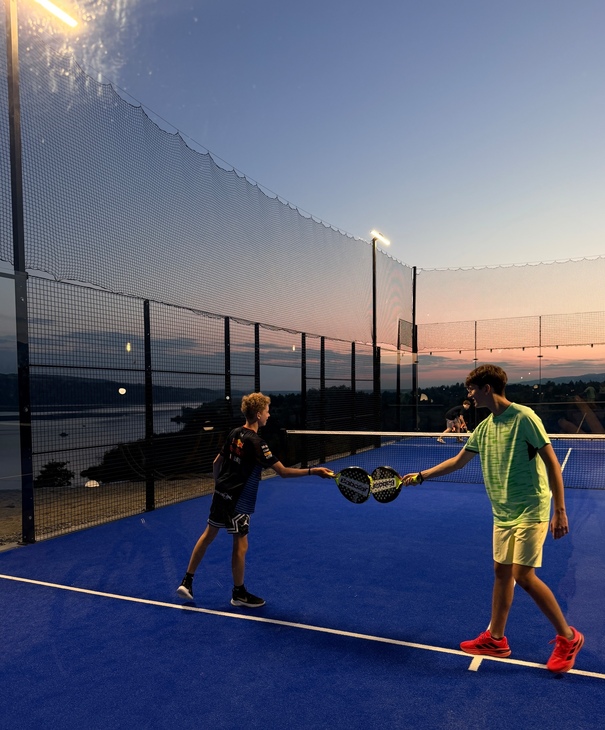 Rooftop illuminated padel court with blue surface and protective netting, teens clinking paddles at sunset over sea view.
