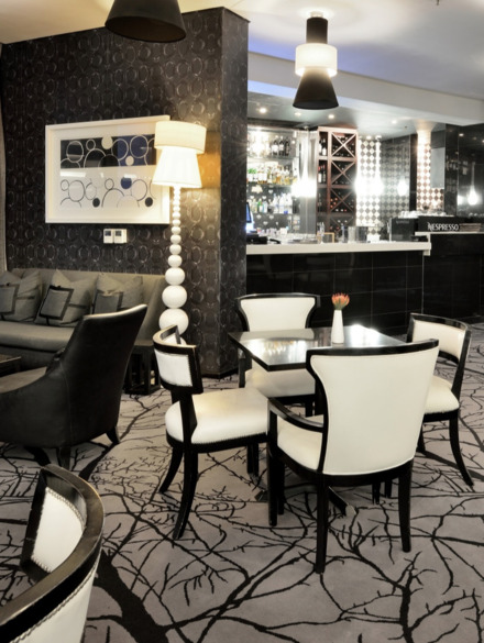 Moderne Hotel-Lounge mit schwarz-weißen Sitzmöbeln, gemustertem Teppich, bodentiefen Fenstern und Pendelleuchten.