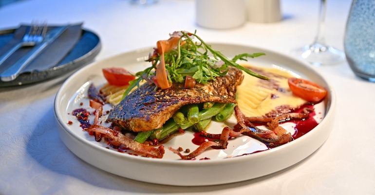 Seared fish fillet on green beans with mashed potatoes, crispy bacon, arugula and red wine glaze on a white tablecloth.
