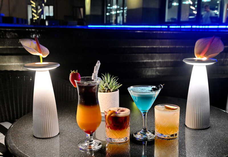 四杯色彩鸡尾酒摆于酒店圆桌，配有发光台灯、观赏多肉植物和蓝色氛围灯，夜晚露台氛围