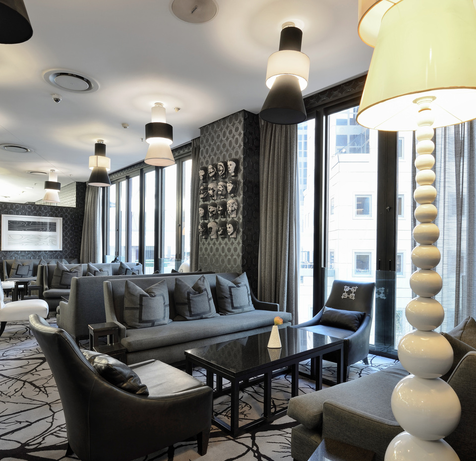 Salón de hotel contemporáneo con sofás grises, sillones de cuero, lámparas colgantes, alfombra con diseño y ventanales con vistas a la ciudad