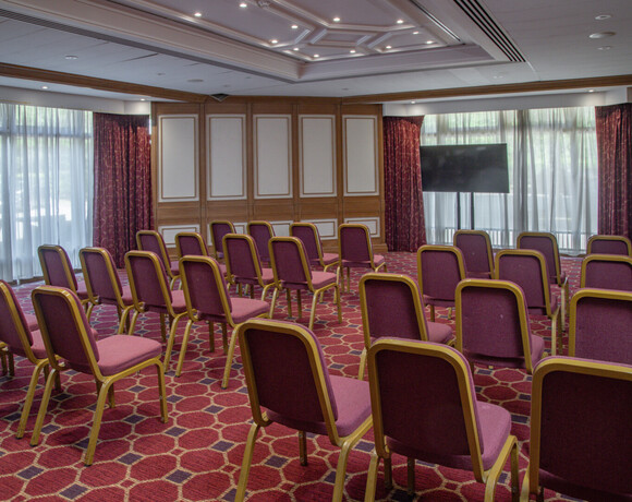 Hotel meeting room with rows of burgundy chairs, patterned carpet, wood-paneled walls, AV screen and sheer curtains.