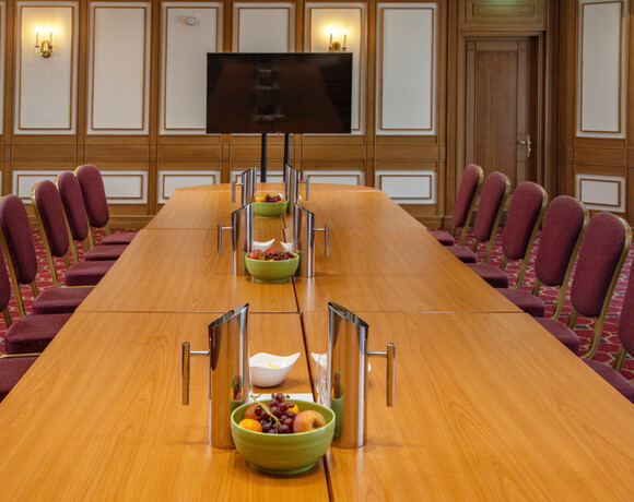 Elegant hotel boardroom with long wood conference table, burgundy chairs, wall-mounted TV, fruit bowls and stainless pitchers.