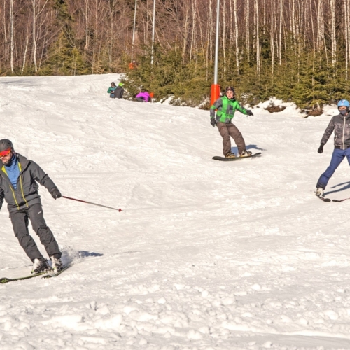 Tři hosté — lyžař a dva snowboardisté v helmách — sjíždějí slunný svah hotelového střediska s březovým lesem v pozadí