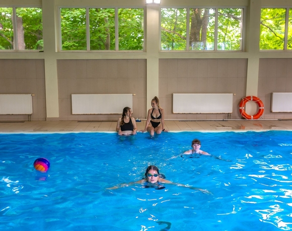 Krytý hotelový bazén s velkými okny, hosté plavou a povídají si, barevný míč pluje vodou a záchranný kruh je u okraje