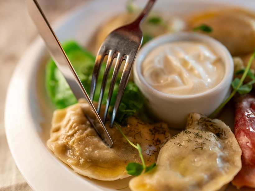Ręka kroi pieroga na talerzu: pierogi podane z kwaśną śmietaną, koperkiem i małą sałatką.