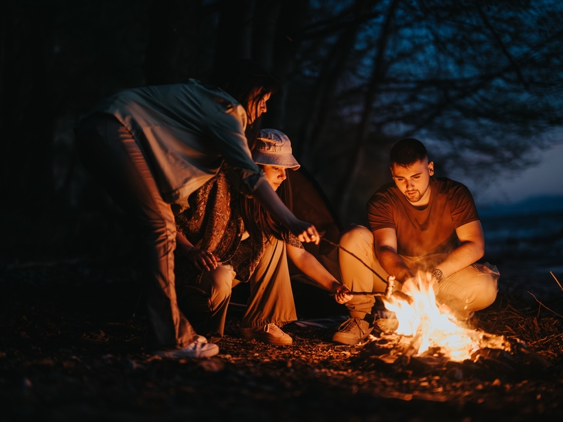 Grupa przy ognisku na kempingu przy brzegu, trzy osoby pieką nad ogniem na patykach, namiot i nocna zieleń w tle
