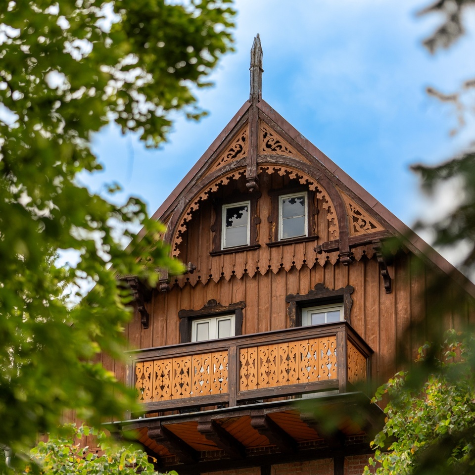 Drewniany pensjonat w stylu alpejskim — ozdobny szczyt z rzeźbionymi detalami i balkonem, widok przez zielone liście.
