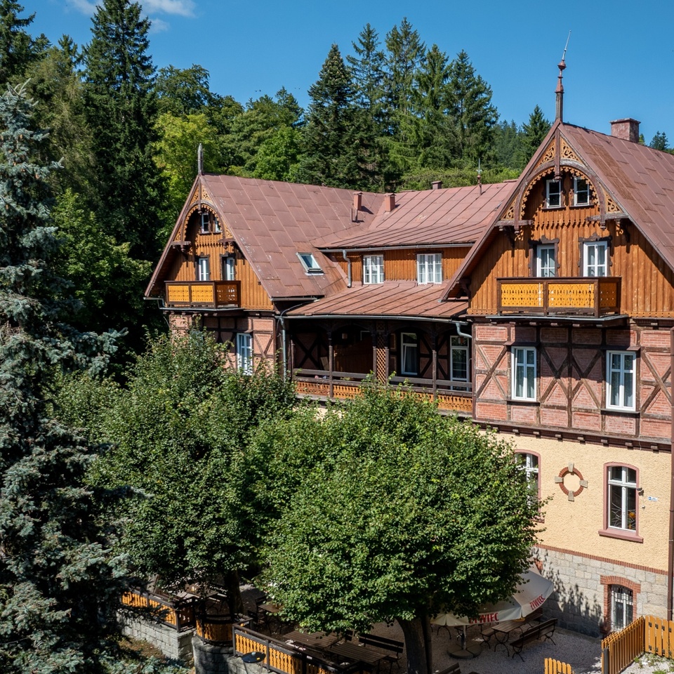 Zabytkowa willa-hotel z drewnianymi balkonami, zewnętrznym tarasem i stolikami pod drzewami, otoczona gęstym lasem