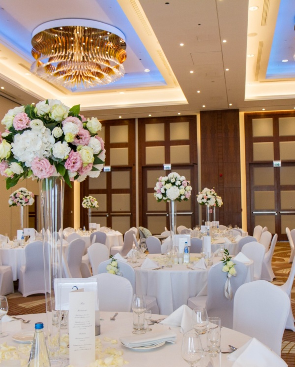 Elegancka sala bankietowa z okrągłymi stołami, białymi obrusami, wysokimi kwiatowymi dekoracjami i złotymi żyrandolami.