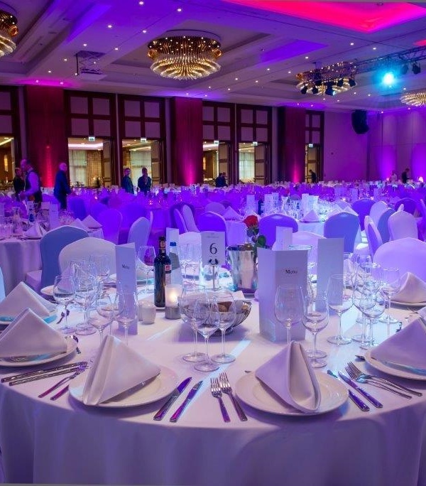 Elegancka sala bankietowa z okrągłymi stołami, białymi obrusami, nastrojowym fioletowym oświetleniem i kryształowymi żyrandolami.