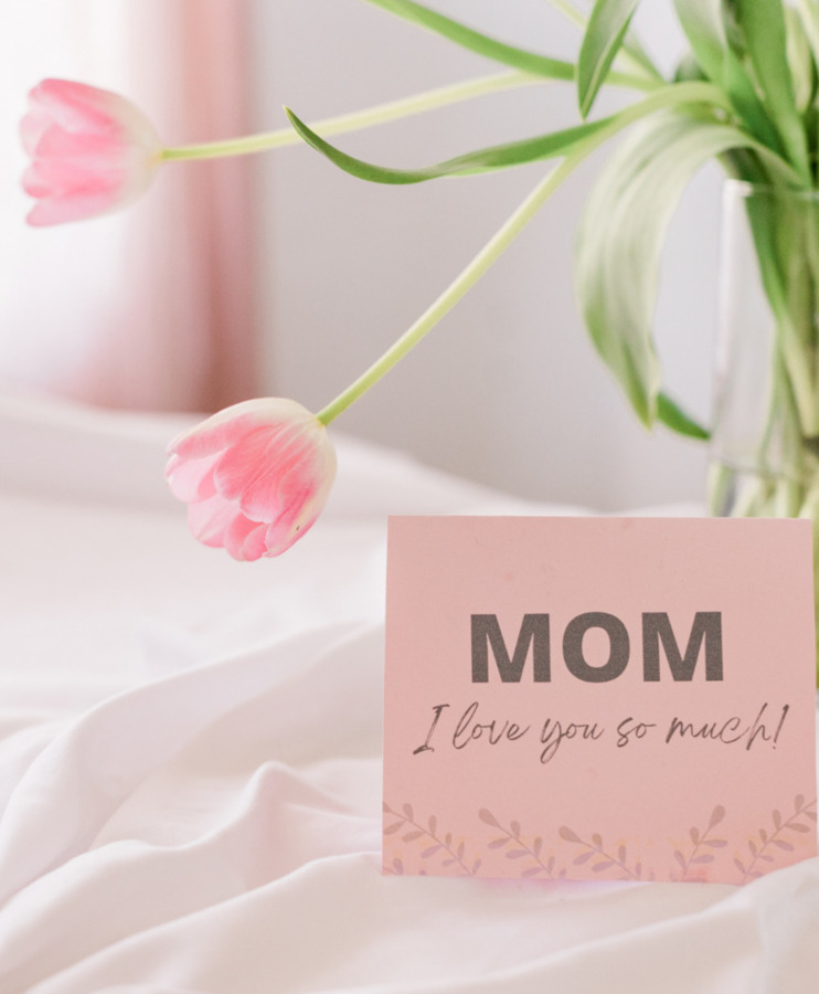 Soft pink tulips in a vase on white bedding next to a card saying "Mom, I love you so much"