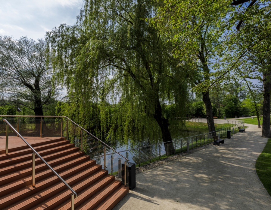 Taras z drewnianymi schodami nad wodą otoczony zielonymi drzewami i ścieżką spacerową w hotelowym ogrodzie