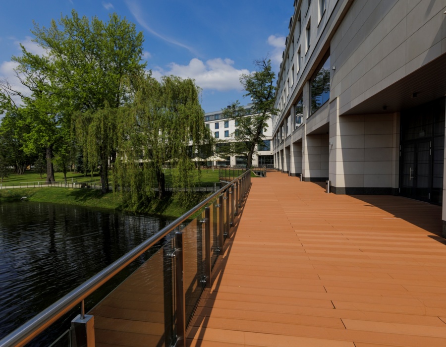 Przestronny taras hotelowy z szklaną balustradą nad stawem i widokiem na zielony park obok budynku.