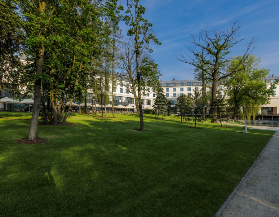 Hotel otoczony dużym, zadbanym trawnikiem z wysokimi drzewami i tarasem restauracyjnym pod parasolami na świeżym powietrzu.