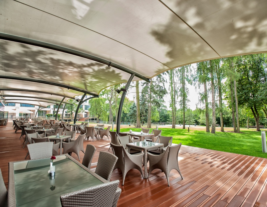 Przestronny, zadaszony taras hotelowej restauracji z wiklinowymi krzesłami i widokiem na zielony ogród z wysokimi drzewami.