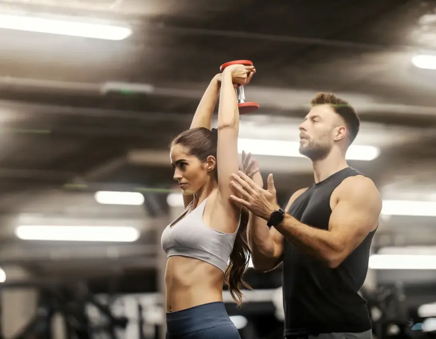 Trener personalny pomaga kobiecie podczas ćwiczenia z hantlem w nowoczesnej hotelowej siłowni z oświetleniem sufitowym.