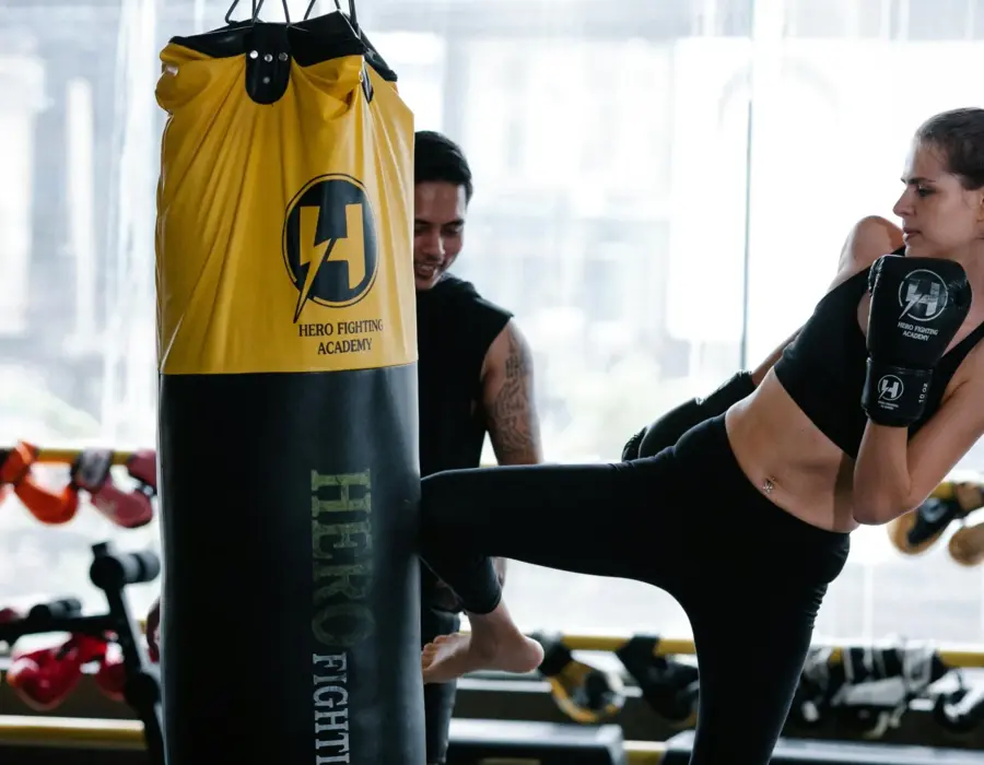 Kobieta trenuje kopnięcie na worek bokserski w nowoczesnym klubie fitness z trenerem w tle