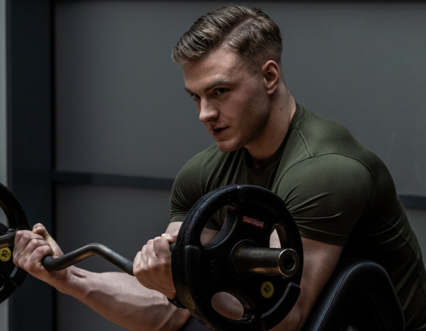 Mężczyzna ćwiczy biceps na siłowni hotelowej, używając sztangi na specjalnej ławce do uginania ramion.