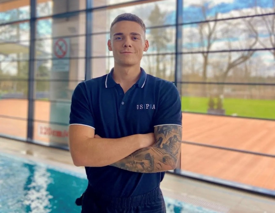 Mężczyzna z tatuażami stoi z założonymi rękami obok basenu w nowoczesnym hotelowym spa z dużymi oknami i widokiem na zieleń.