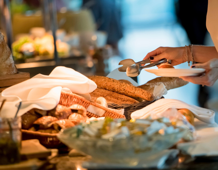 Fresh bread and baguettes displayed at hotel breakfast buffet, guest using tongs to serve onto plate.
