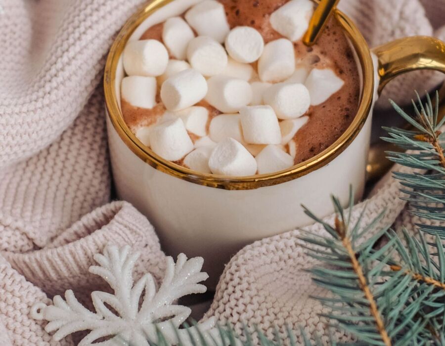 Kubek gorącej czekolady z piankami na ciepłym, plecionym kocu, ozdobiony świąteczną gałązką i białą dekoracją śnieżynki