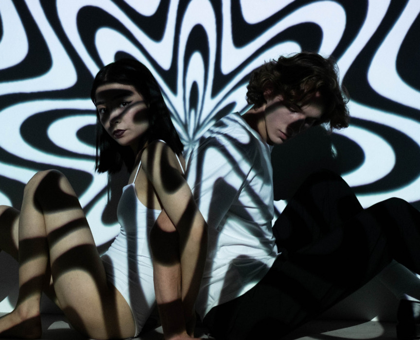 Dwoje młodych ludzi siedzi oparci plecami na tle dynamicznych, czarno-białych wzorów świetlnych tworzących psychodeliczny efekt.