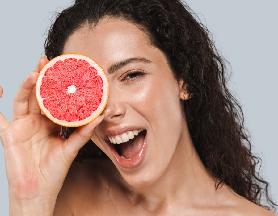 Woman with glowing skin holding half a grapefruit over her eye, smiling, radiant and natural