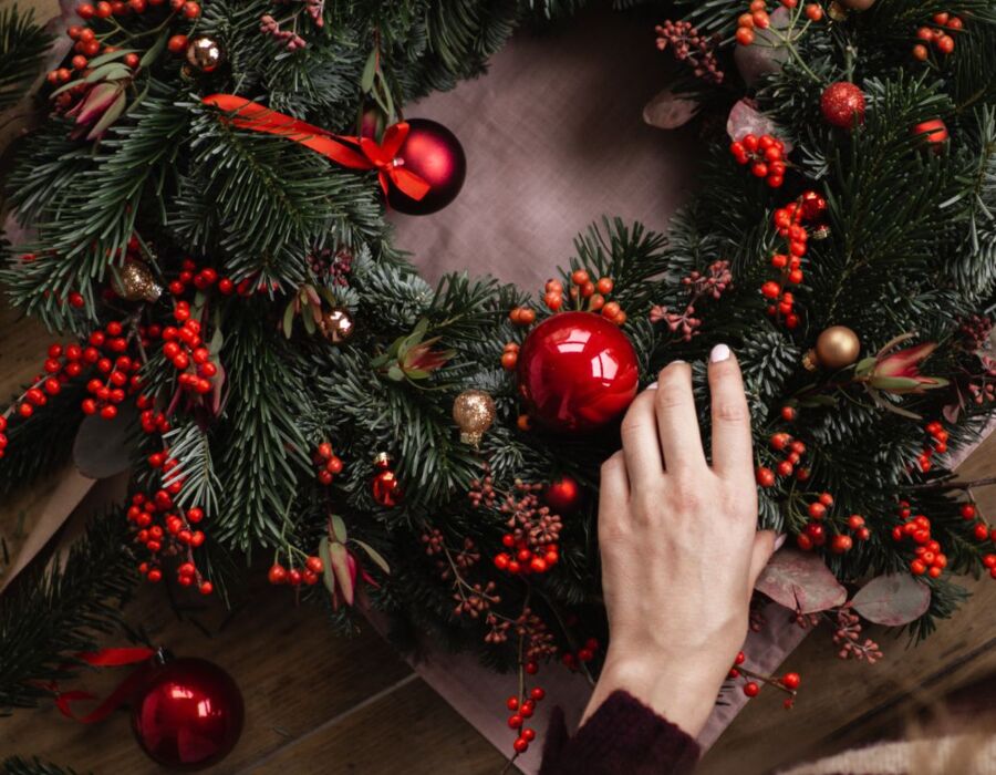 Ręka dekorująca świąteczny wieniec z zielonych gałązek, czerwonych jagód i bombek na drewnianym stole