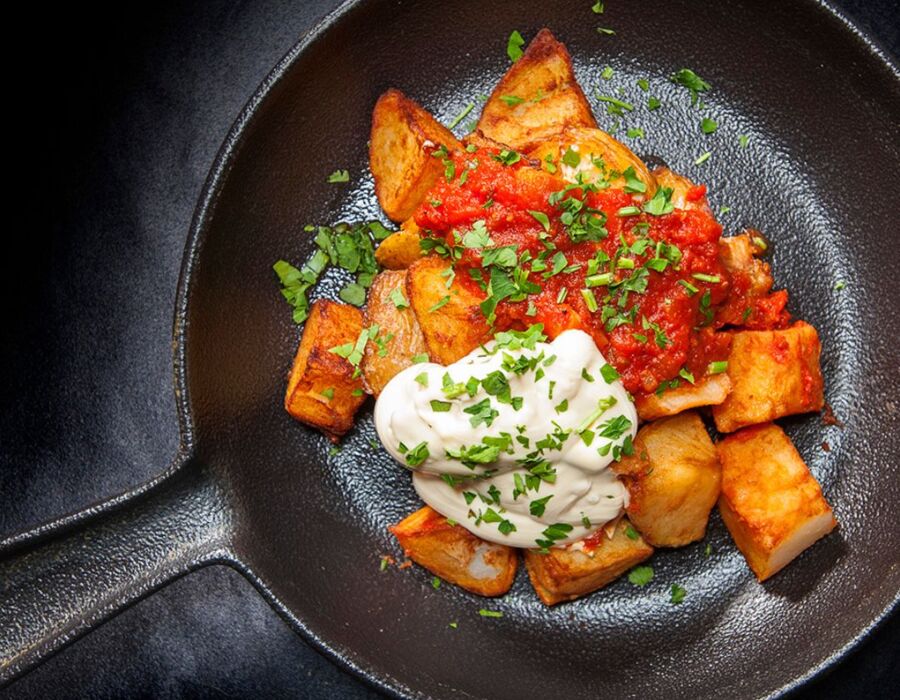 Kawałki smażonych ziemniaków podane na czarnym talerzu z czerwonym sosem i kleksem śmietany, posypane świeżą natką pietruszki