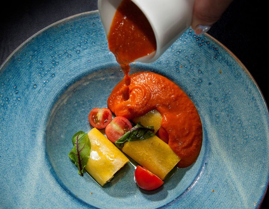 Ręka wylewająca gęsty czerwony sos na ravioli z pomidorami i zielonymi liśćmi na niebieskim talerzu