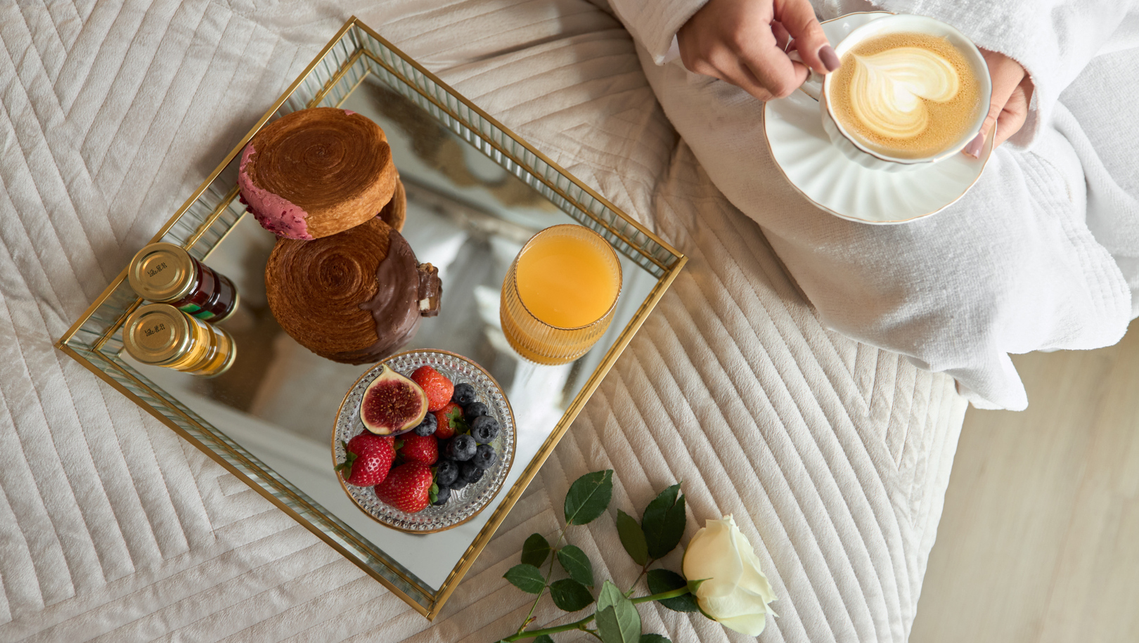 Lustrzana taca ze śniadaniem na miękkim łóżku: ciastka, świeże owoce, sok pomarańczowy i latte w dłoniach gościa w białym szlafroku.