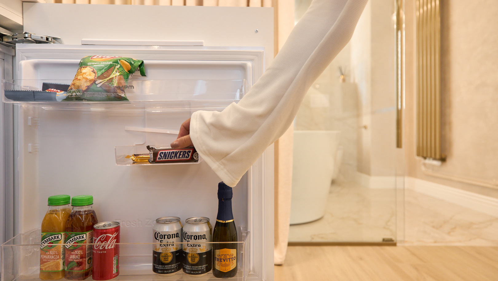 Minibar przy łazience w pokoju z piwami, sokami, mini prosecco i przekąskami; ręka gościa sięga po Snickersa